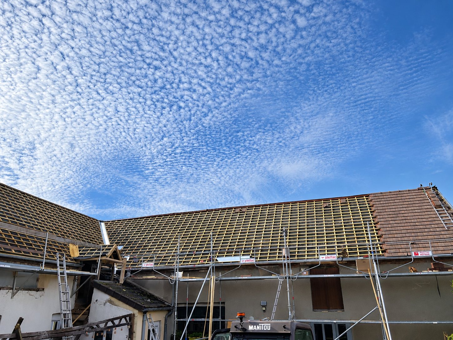 Chantier de rénovation d'une toiture dont les tuiles sont retirées, entourée d'un échafaudage, avec un engin Manitou au premier plan sous un ciel moutonné.
