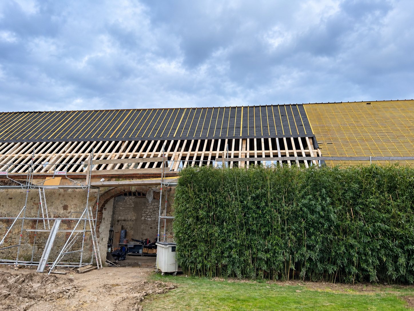 Chantier de rénovation d'une grande toiture de grange en pierre, avec liteaux et écran noir posés, échafaudage au premier plan et haie de bambous à droite.