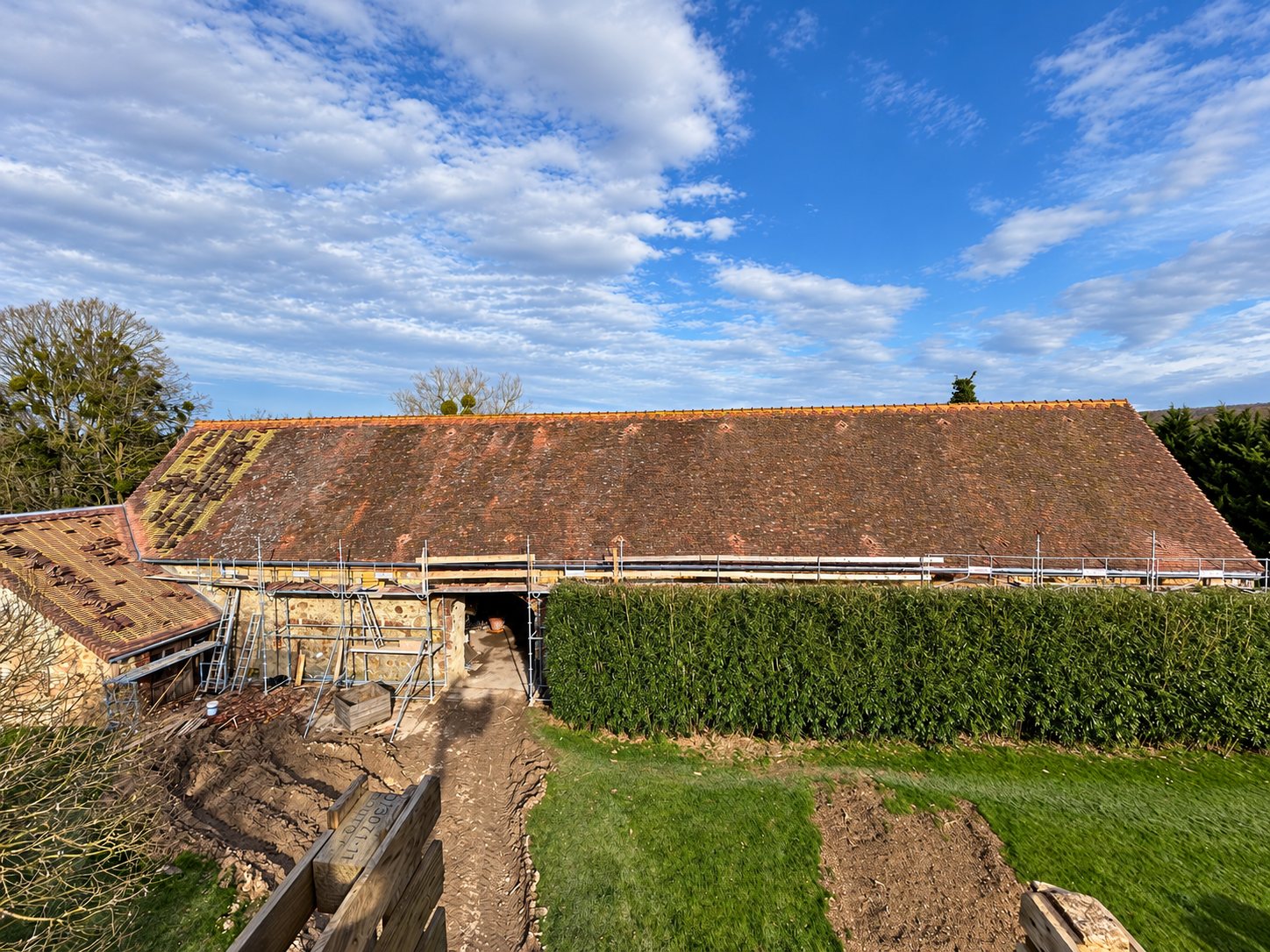 Une longue grange en pierre à toit de tuiles anciennes, en cours de rénovation avec un échafaudage, une partie des tuiles retirées et de la terre remuée au premier plan.