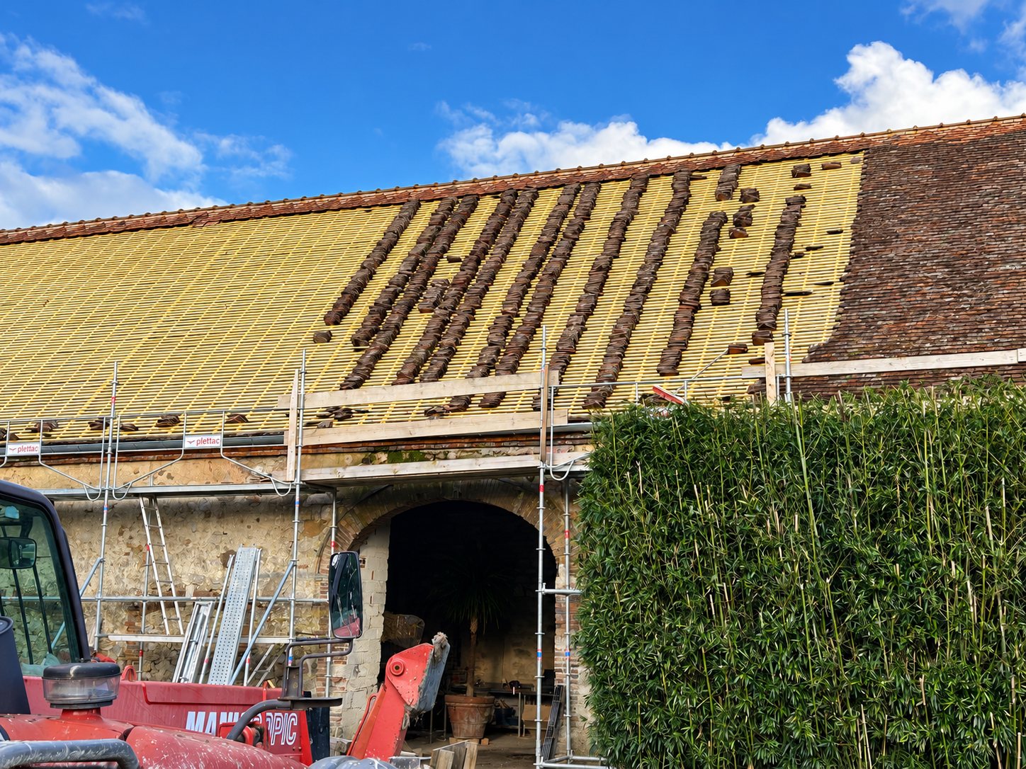 Chantier de rénovation d'un toit de grange dont les tuiles sont partiellement déposées, entouré d'un échafaudage et d'un chariot télescopique rouge.