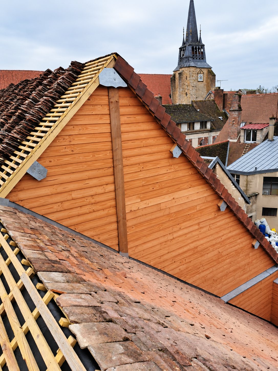Toiture en cours de rénovation avec un pignon recouvert de lames de bois, des tuiles anciennes et un clocher de village en arrière-plan.