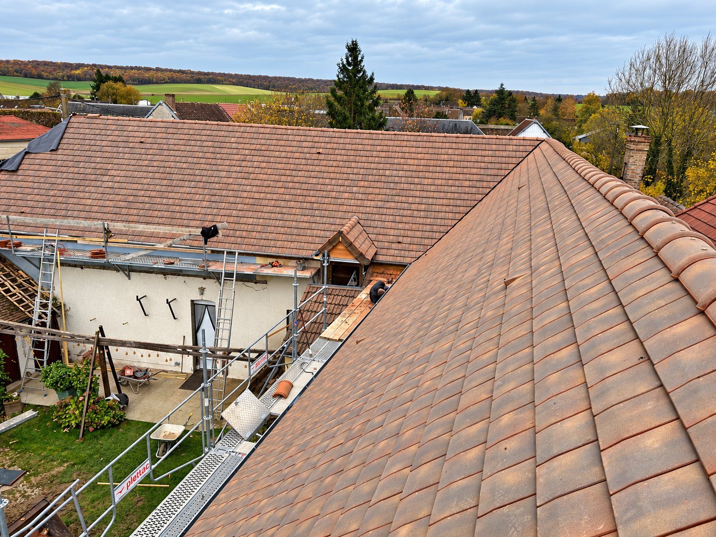 Vue en hauteur d'un chantier de réfection de toiture en tuiles rouges avec échafaudage et ouvriers, dans un village entouré de champs.