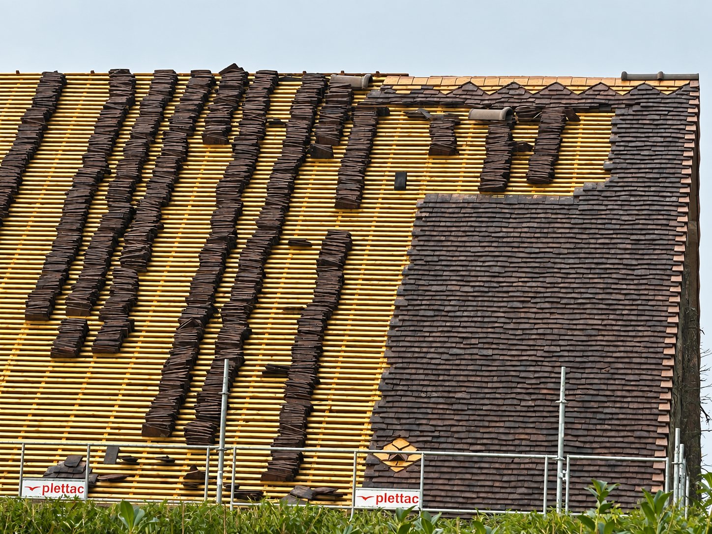Un grand toit en cours de rénovation dont une partie des tuiles plates brunes a été déposée et empilée sur les lattes en bois, protégé par un garde-corps d'échafaudage.