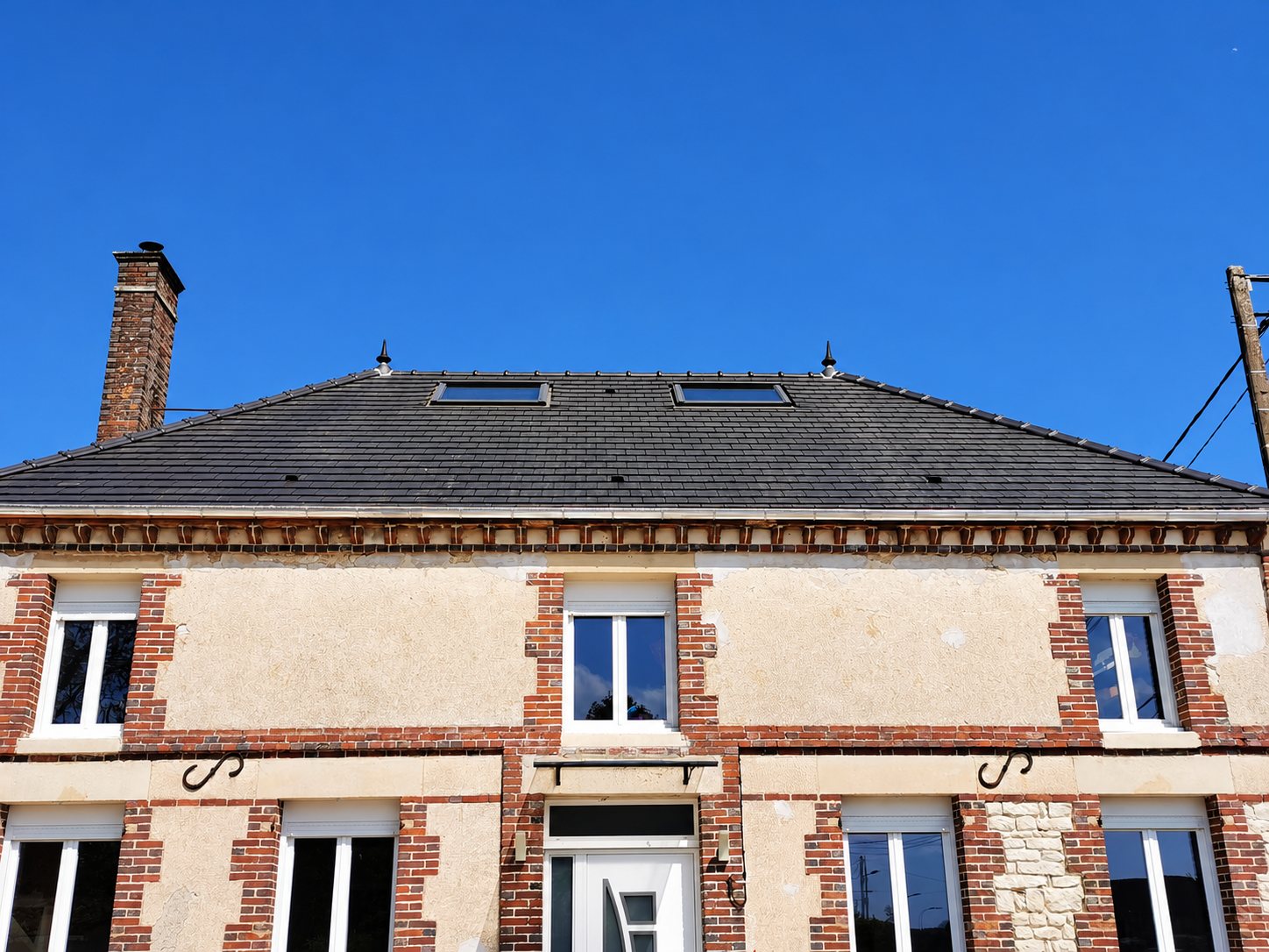 Façade d'une maison ancienne en briques et pierres dont la toiture en ardoise neuve est équipée de deux fenêtres de toit, sous un ciel bleu.