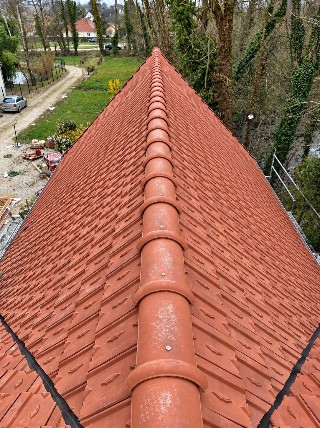 Vue en hauteur d'un toit fraîchement posé en tuiles de terre cuite rouge avec son faîtage, au-dessus d'un jardin et d'un chantier.