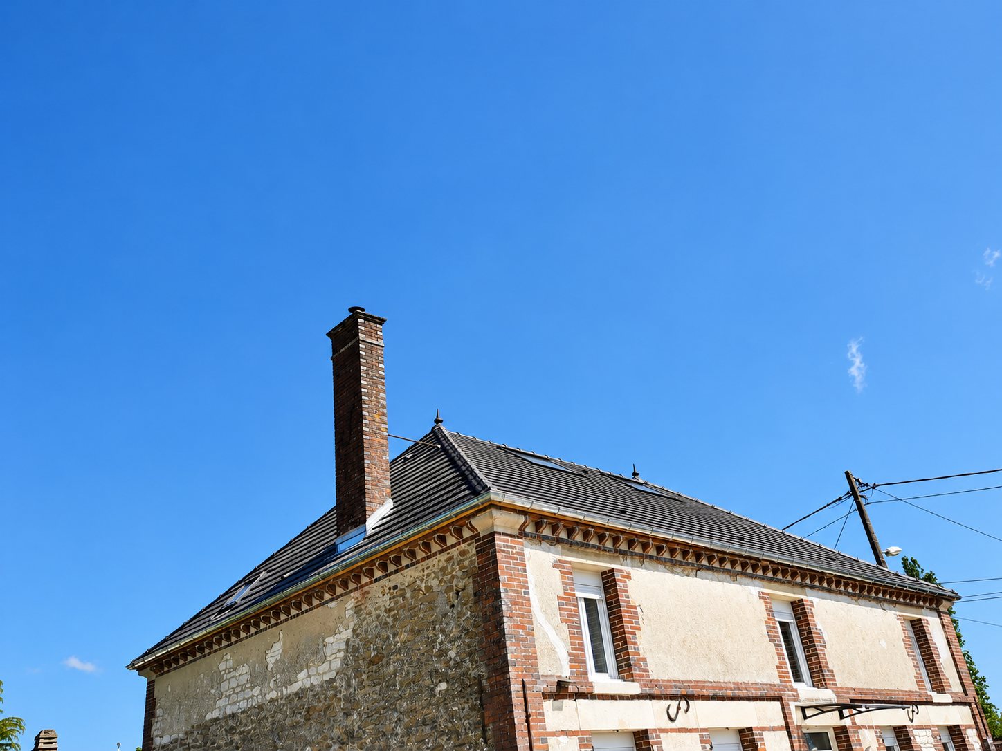 Maison ancienne en brique et pierre avec une toiture en ardoise récemment rénovée et une haute cheminée en brique, sous un ciel bleu dégagé.