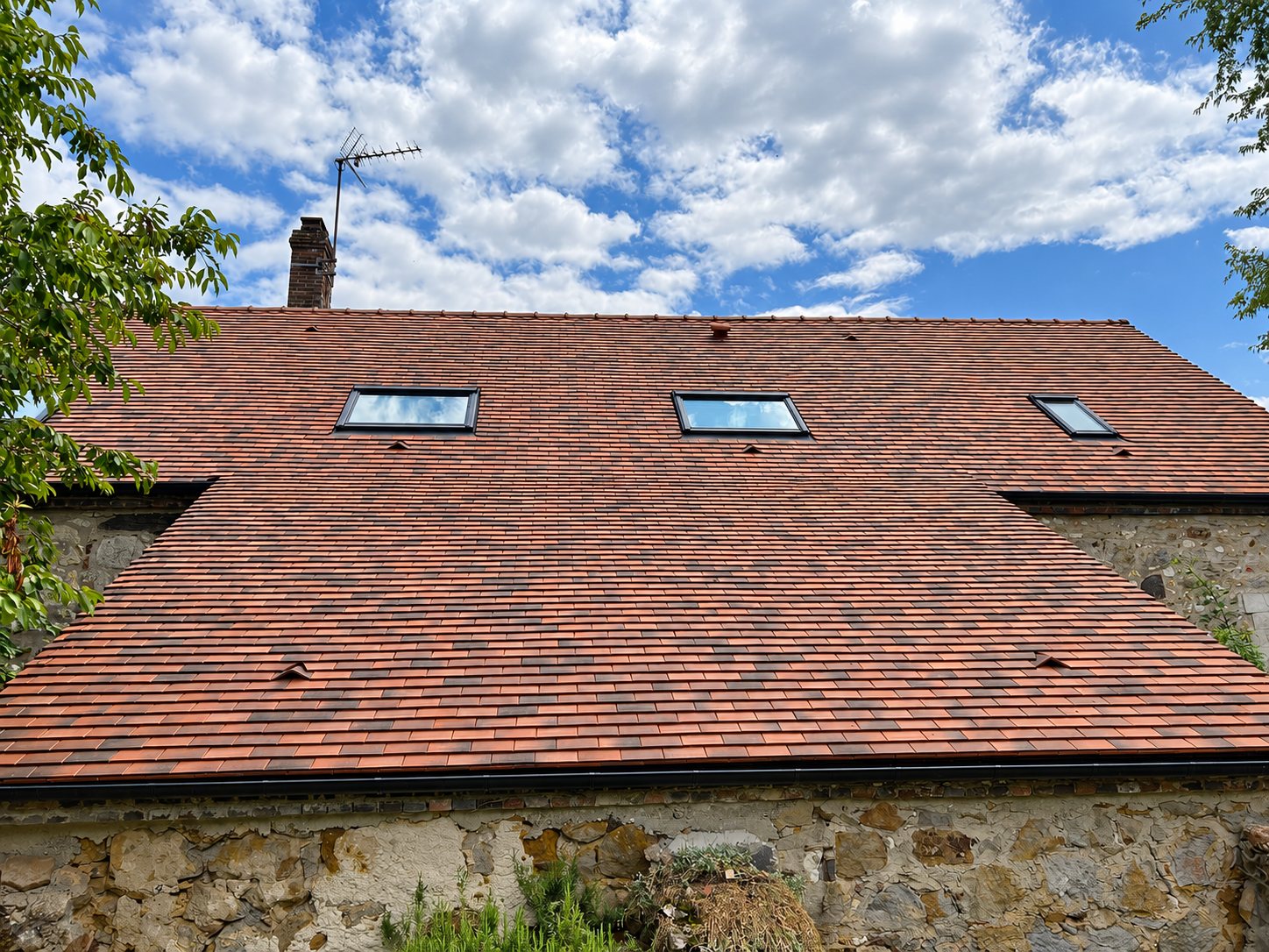 Vue d'une toiture rénovée en petites tuiles plates rouges et brunes, équipée de trois fenêtres de toit, au-dessus d'un mur en pierre sous un ciel nuageux.