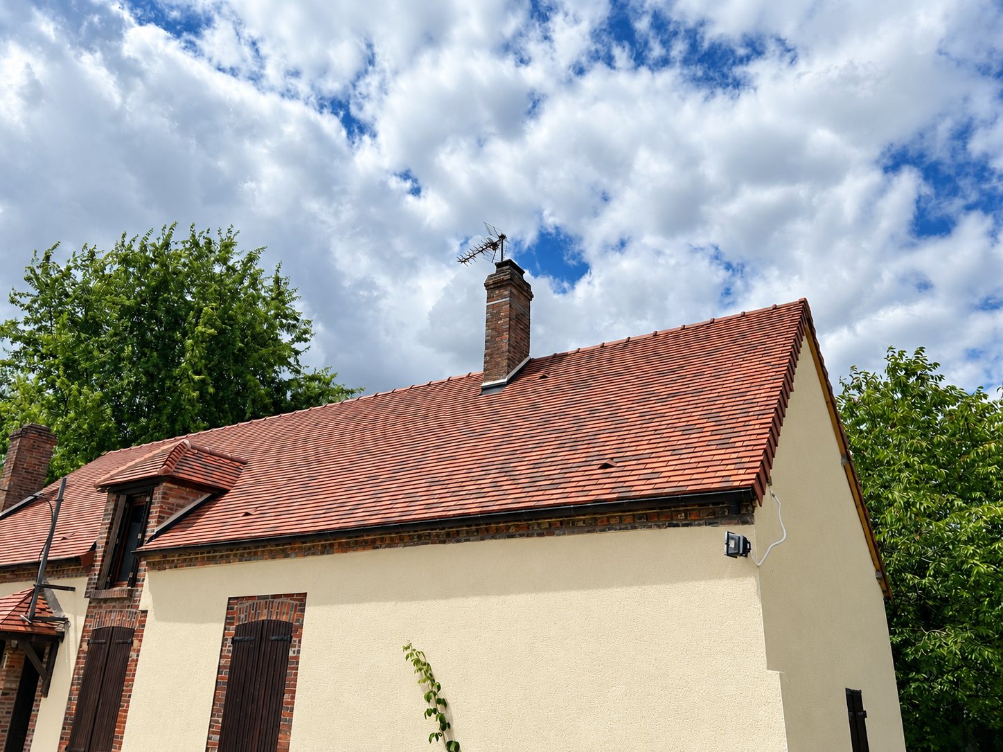 Toit récemment rénové en tuiles plates rouge nuancé avec cheminée en briques et antenne, sur une maison aux murs crépis beiges, sous un ciel nuageux.
