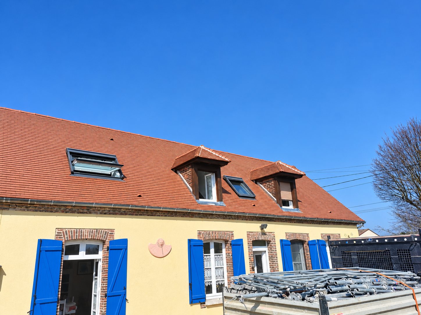 Maison à façade jaune et volets bleus dont la toiture en tuiles rouges est équipée de lucarnes et de fenêtres de toit, avec des échafaudages empilés au premier plan.